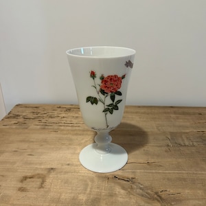 May include: White porcelain goblet with a floral design featuring a pink rose with green leaves and a small bud. The goblet has a stem and a round base. The text "Rosa Gallica Officinalis" is printed on the goblet.