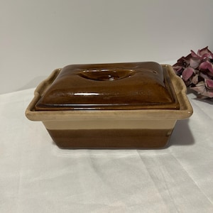 Peut inclure: Un plat à casserole rectangulaire en céramique avec un couvercle. Le plat est bicolore, avec une base beige clair et un dessus et un couvercle marron foncé. Le couvercle a une petite poignée. Le plat est sur une surface blanche.