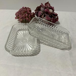 Puede incluir: Dos platos rectangulares de vidrio transparente con un diseño geométrico texturizado. Los platos están colocados sobre una superficie blanca, con flores moradas secas en el fondo. Los platos tienen un patrón de rayos de sol en el centro.