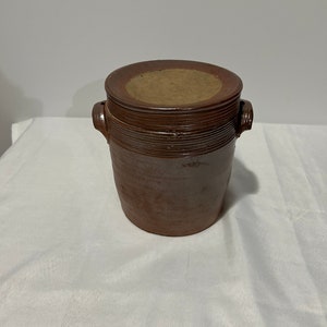 K&ouml;nnte beinhalten: Ein brauner Keramiktopf mit Deckel. Der Topf hat zwei Henkel und eine leicht abgerundete Form.