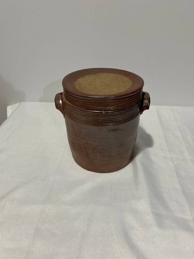K&ouml;nnte beinhalten: Ein brauner Keramiktopf mit Deckel. Der Topf hat zwei Henkel und eine leicht abgerundete Form.