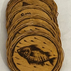 May include: A set of six wooden coasters with a woven wicker rim. Each coaster features a different design, including a fish, a bird, and a floral pattern. The coasters are a natural wood color with a dark brown stain.