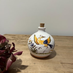May include: A white ceramic bottle with a cork stopper, featuring a hand-painted bird design in yellow, blue, and orange. The bottle has a round shape and is decorated with floral accents. A decorative item for the home.