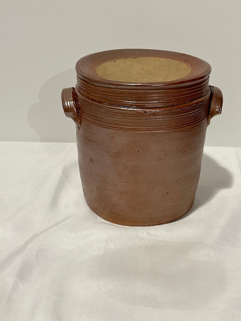 K&ouml;nnte beinhalten: Ein brauner Steinzeugtopf mit Deckel. Der Topf hat einen runden K&ouml;rper und zwei Henkel. Der Deckel ist aus einem helleren braunen Material.