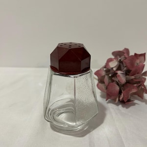 May include: A clear glass salt shaker with a textured surface and a faceted, dark red lid. The lid has several small holes. The shaker is empty and sits on a white surface. A dried flower arrangement is in the background.