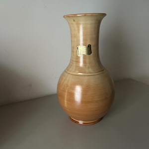 Puede incluir: Un jarrón de cerámica marrón claro con cuello estrecho y base ancha. El jarrón tiene una etiqueta en el lateral que dice "Hecho a mano en los EE. UU.".