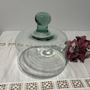 Peut inclure: Une cloche en verre transparent avec un sommet arrondi et une poignée sphérique de couleur vert d'eau. La cloche présente une base circulaire large et un corps transparent, conçue pour couvrir des objets tout en restant visible.