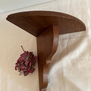 May include: A wooden wall shelf with a curved top and a decorative, carved bracket. The shelf is made of dark brown wood and has a rustic appearance.