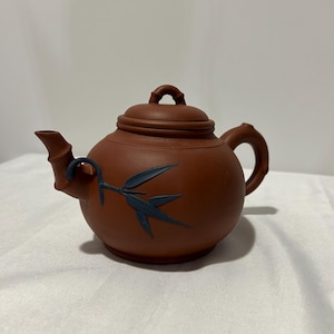 Peut inclure: Une théière en céramique brune avec un motif de bambou. La théière a un couvercle avec une petite poignée et un bec verseur avec une poignée en bambou. La théière est posée sur une surface blanche.