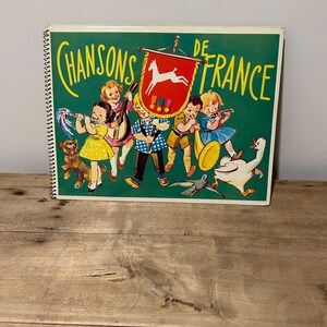 May include: A vintage children's book titled "Chansons de France" with a green cover featuring a colourful illustration of children playing musical instruments. The cover also includes a white horse on a red banner with gold trim.