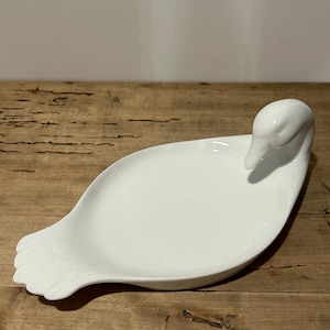 Op de afbeelding: Een witte keramische schaal in de vorm van een zwaan met een lange, gebogen nek en een breed, plat lichaam. De schaal is perfect om kleine snacks te serveren of om sieraden in te bewaren.