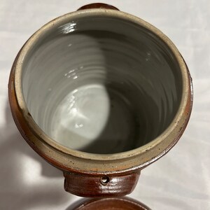 K&ouml;nnte beinhalten: Eine braune Keramik-Dose mit Deckel. Die Dose hat zwei Griffe und ein kleines Loch an der Seite. Der Deckel ist ebenfalls braun und hat ein kleines Loch in der Mitte.