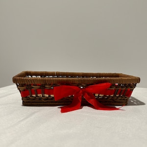 Op de afbeelding: Een bruine rieten mand met een rode strik. De mand is rechthoekig en heeft een geweven ontwerp.