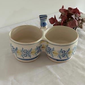 Peut inclure: Un double bol en céramique avec une poignée, de couleur crème et orné de motifs floraux bleus et jaunes. Le bord et la base sont soulignés en bleu. Une pièce décorative pour le service.