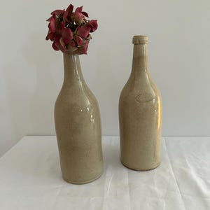Könnte beinhalten: Zwei Keramikflaschen, eine mit einem Trockenblumenarrangement, beide in hellbeige.