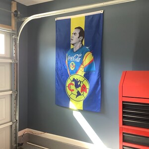 May include: A blue and yellow flag with a portrait of a man wearing a soccer jersey with the word "Coca-Cola" on it. The flag also features a soccer ball with the letters "CCA" on it.