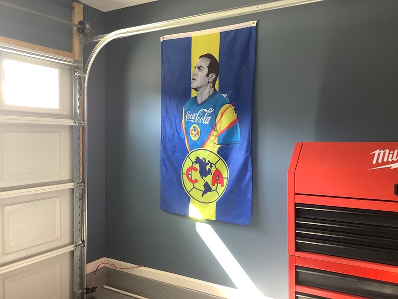 May include: A blue and yellow flag with a portrait of a man wearing a soccer jersey with the word "Coca-Cola" on it. The flag also features a soccer ball with the letters "CCA" on it.