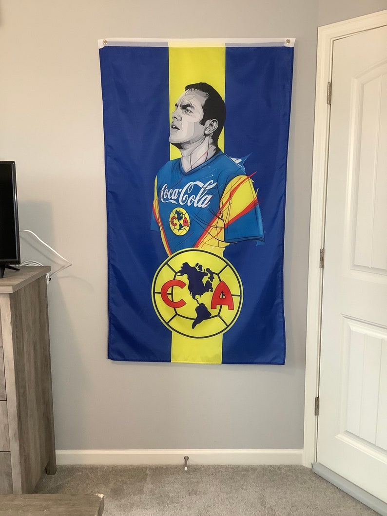 May include: A blue and yellow flag with a soccer player wearing a blue jersey with the text "Coca-Cola" on the front. The player is looking up and has a yellow and red stripe on his jersey. The flag also has a yellow circle with a soccer ball and the letters "C" and "A" in red.