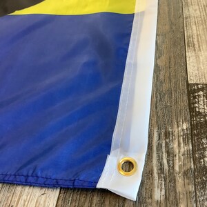 May include: Close-up of a blue and yellow flag with a white border and a brass grommet. The flag is made of fabric and has a stitched seam.