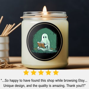 May include: A lit candle in a glass jar with a black label featuring a cartoon ghost reading a book. The ghost is white, the book is brown, and the background is green. The candle is on a wooden surface.
