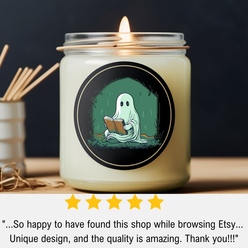 May include: A lit candle in a glass jar with a black label featuring a cartoon ghost reading a book. The ghost is white, the book is brown, and the background is green. The candle is on a wooden surface.
