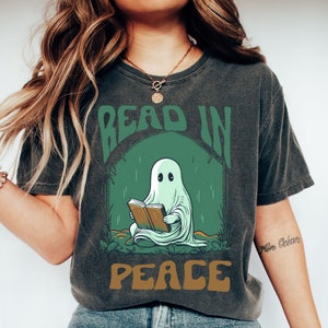 May include: Dark gray t-shirt with a graphic of a ghost reading a book inside a green arch. The words "READ IN PEACE" are printed above and below the ghost illustration in green and brown lettering.