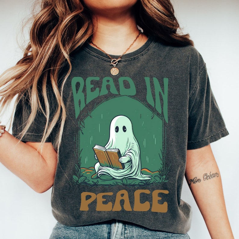 May include: Dark gray t-shirt with a graphic of a ghost reading a book inside a green arch. The words "READ IN PEACE" are printed above and below the ghost illustration in green and brown lettering.