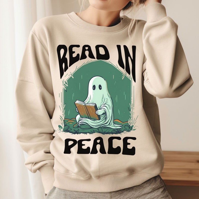 May include: Beige sweatshirt with a graphic of a ghost reading a book. The ghost is light green and is sitting in a green archway. The words "READ IN PEACE" are printed above and below the ghost in black.