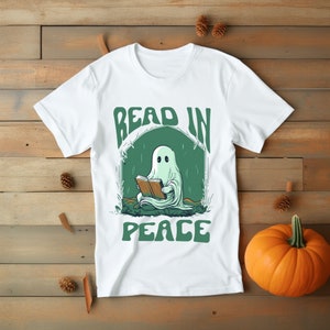 May include: White t-shirt with a green and white graphic of a ghost reading a book. The text "READ IN PEACE" is above and below the ghost illustration. The shirt is displayed on a wooden surface with pinecones and a pumpkin.