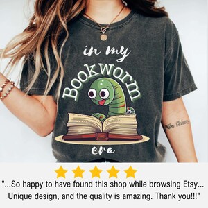May include: Dark gray t-shirt featuring a cartoon bookworm peeking over an open book. The text "in my bookworm era" is arched above the worm. The design is in green, white, and brown. The shirt is a casual style.