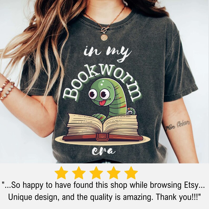 May include: Dark gray t-shirt featuring a cartoon bookworm peeking over an open book. The text "in my bookworm era" is arched above the worm. The design is in green, white, and brown. The shirt is a casual style.