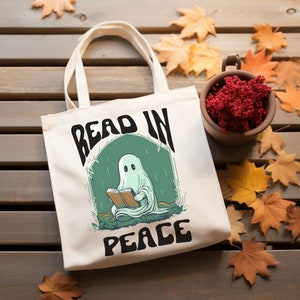 May include: Off-white canvas tote bag with the words "READ IN PEACE" in black. The graphic features a cartoon ghost reading a book inside a green arch. The bag has two off-white handles and is sitting on a wooden surface.