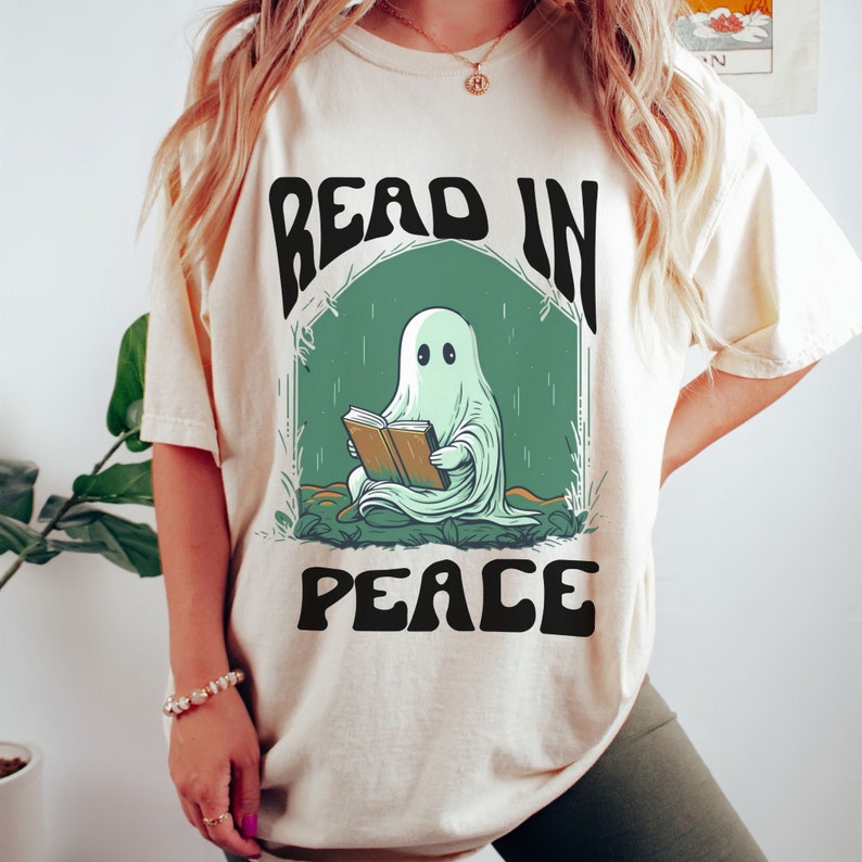 A ghost reading a book with the text 'read in peace' written on a halloween themed tshirt Ivory color. Female model has brown hair, is wearing an oversized tshirt, and is standing in front of a potted plant.