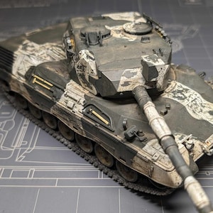 Puede incluir: Un modelo detallado de un tanque militar con un esquema de pintura de camuflaje en negro, blanco y verde. El tanque tiene un cañón largo y una torreta con una escotilla abierta. El tanque está sentado sobre una superficie gris.