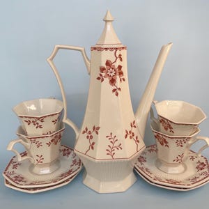 May include: A cream-colored porcelain coffee set with a tall, geometric coffee pot and four cups and saucers. The set is decorated with a red floral pattern. The cups and saucers are stacked in pairs.