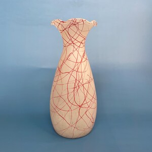 May include: A tall, off-white ceramic vase with a flared, ruffled rim. The vase is adorned with a random pattern of red lines. The vase has a rounded base and tapers upwards, creating an elegant silhouette. Home decor.