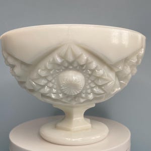 May include: A white, footed bowl with intricate, raised geometric patterns. The bowl has a wide, rounded rim and a pedestal base. The design includes diamond and starburst motifs. The bowl is made of a glossy, opaque material.