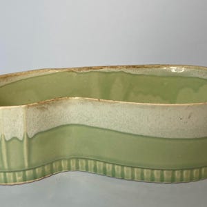 May include: A green ceramic planter with a wavy, asymmetrical shape. The planter has a white band around the middle and a textured, ribbed base.