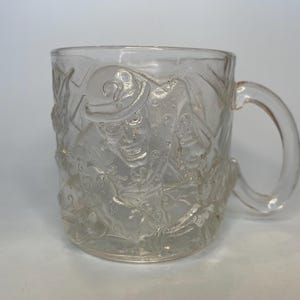 May include: Clear glass mug with a detailed embossed design featuring a figure in a hat. The mug has a rounded handle and a cylindrical shape. The design is visible on the exterior of the mug.