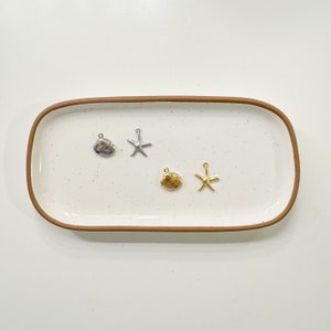 May include: Four silver and gold charms on a white ceramic tray. The charms are a seashell, a starfish, a seashell, and a starfish.