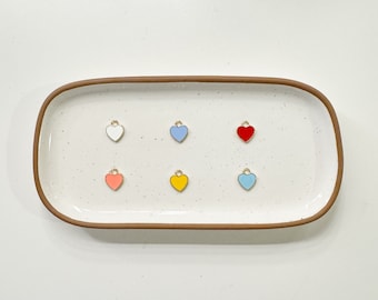 Puede incluir: Un conjunto de seis dijes en forma de corazón en varios colores, incluyendo blanco, azul claro, amarillo, rosa, rojo y azul claro. Los dijes están dispuestos en dos filas de tres en una bandeja de cerámica blanca con un borde marrón.
