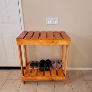 Puede incluir: Un zapatero de madera con dos estantes. El estante superior está hecho de listones de madera paralelos. El estante inferior contiene varios pares de zapatos. El estante es de madera marrón claro y está colocado contra una pared beige.