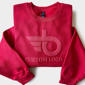 May include: A folded red sweatshirt with a crew neck and long sleeves. The sweatshirt features a tonal custom logo design on the front. The text "CUSTOM LOGO" is below the logo. The garment is made of a soft material.