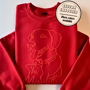 May include: A red sweatshirt with a custom embossed dog design. The design features a dog with its tongue out and the text "The Maristottis". A circular sticker in the upper right corner says "Custom Embossed" and "More colors available".