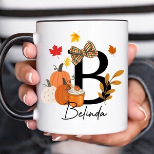 May include: White ceramic mug with a black rim and handle, featuring a fall-themed design. The design includes pumpkins, fall leaves, a bow, and the letter "B" with the name "Belinda" in script. The mug is held by a person.