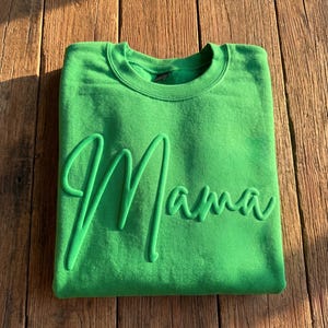 May include: A bright green sweatshirt with the word "Mama" in a raised, cursive font. The sweatshirt is folded neatly on a wooden surface, with the word "Mama" prominently displayed across the front.