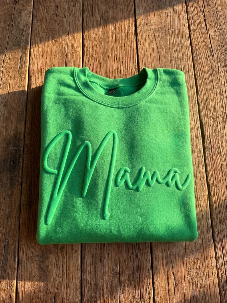 May include: A bright green sweatshirt with the word "Mama" in a raised, cursive font. The sweatshirt is folded neatly on a wooden surface, with the word "Mama" prominently displayed across the front.