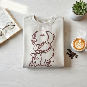 May include: Beige sweatshirt with a brown outline of a dog and the text "The Marciottis". The image also includes a cup of coffee with latte art, a succulent plant, a book, and eyeglasses.
