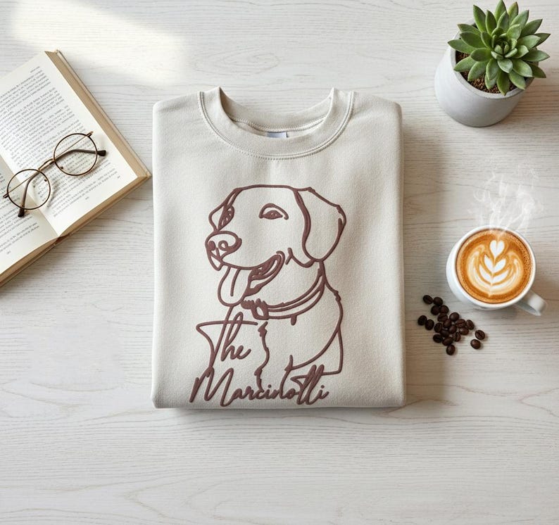 May include: Beige sweatshirt with a brown outline of a dog and the text "The Marciottis". The image also includes a cup of coffee with latte art, a succulent plant, a book, and eyeglasses.
