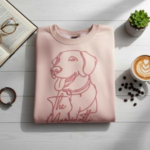 May include: A light pink sweatshirt with a dog graphic and the text "The Marsiliotti". The sweatshirt is next to a cup of coffee, a succulent plant, an open book, and a bracelet.